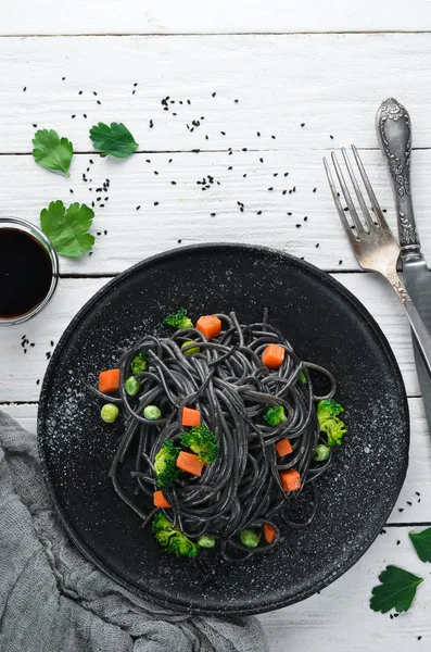 Black pasta with vegetables on a black plate. Top view. On a white wooden background. Free copy space