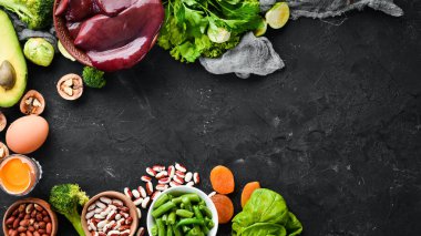 Doğal B9 vitamini içeren gıdalar: Karaciğer, avokado, brokoli, ıspanak, maydanoz, fasulye, fındık, siyah taş arka plan üzerinde. Üstte görüntü.