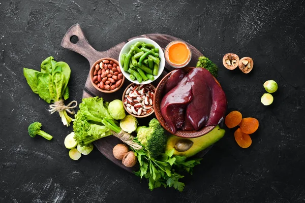 Doğal B9 vitamini içeren gıdalar: Karaciğer, avokado, brokoli, ıspanak, maydanoz, fasulye, fındık, siyah taş arka plan üzerinde. Üstte görüntü.