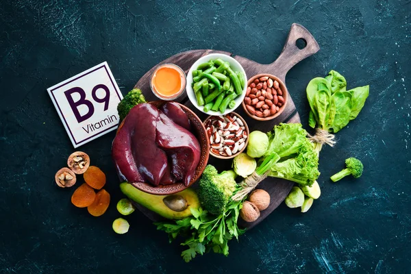 Foods that contain natural vitamin B9: Liver, avocado, broccoli, spinach, parsley, beans, nuts, on a black stone background. Top view.