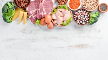 Masada protein oranı yüksek olan yiyecekler. Et, tavuk filetosu, brokoli, fasulye, peynir, yumurta, buğday. Beyaz ahşap arka planda.