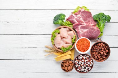 Masada protein oranı yüksek olan yiyecekler. Et, tavuk filetosu, brokoli, fasulye, peynir, yumurta, buğday. Beyaz ahşap arka planda.