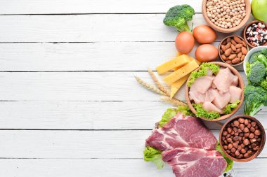 Masada protein oranı yüksek olan yiyecekler. Et, tavuk filetosu, brokoli, fasulye, peynir, yumurta, buğday. Beyaz ahşap arka planda.