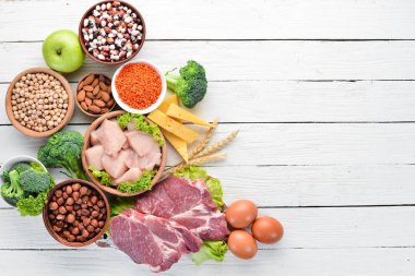 Masada protein oranı yüksek olan yiyecekler. Et, tavuk filetosu, brokoli, fasulye, peynir, yumurta, buğday. Beyaz ahşap arka planda.
