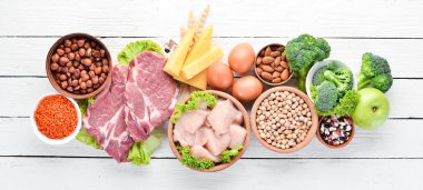 Masada protein oranı yüksek olan yiyecekler. Et, tavuk filetosu, brokoli, fasulye, peynir, yumurta, buğday. Beyaz ahşap arka planda.
