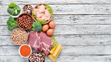 Masada protein oranı yüksek olan yiyecekler. Et, tavuk filetosu, brokoli, fasulye, peynir, yumurta, buğday. Beyaz ahşap arka planda.