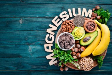 Doğal magnezyum içeren gıdalar. Mg: Çikolata, muz, kakao, fındık, avokado, brokoli, badem. Üst te. Mavi ahşap arka planda.