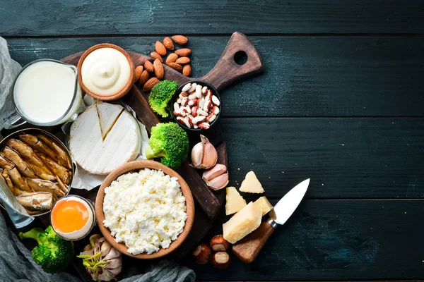 Doğal kalsiyum içeren ürünler: peynir, süt, parmesan, ekşi krema, balık, badem, maydanoz, sarımsak, brokoli. Siyah ahşap bir arka plan üzerinde. Üst te. Ücretsiz kopyalama alanı.