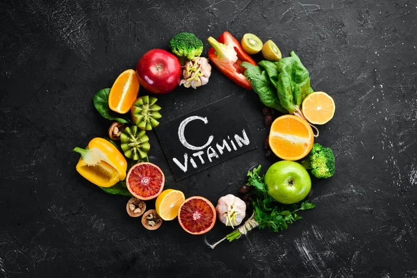 Fruits and vegetables that contain vitamin C: Orange, lemon, apple, roses, garlic, broccoli, apple, kiwi, spinach. Top view. On a black stone background.