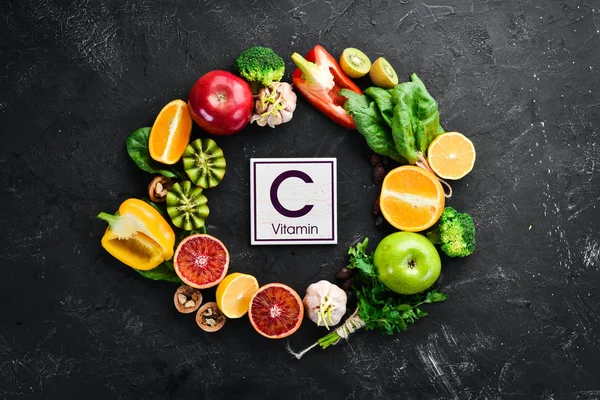 Fruits and vegetables that contain vitamin C: Orange, lemon, apple, roses, garlic, broccoli, apple, kiwi, spinach. Top view. On a black stone background.