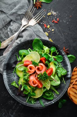 Somon, avokado, domates ve ıspanak salatası. Ahşap arka plan üst görünümünde siyah bir tabak ta. Metniniz için boş alan. Düz yatıyordu
