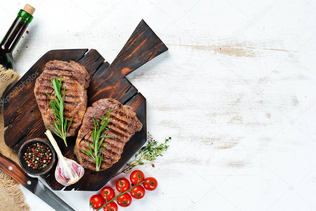 Grilled ribeye beef steak, herbs and spices on a white wooden background. Top view. Free space for your text.