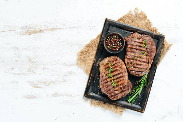 Grilled ribeye beef steak, herbs and spices on a white wooden background. Top view. Free space for your text.