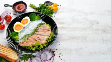 Beyaz ahşap arka planda siyah tabakta tavuk fileto, yumurta ve brokoli ile pirinç eriştesi. Üst Manzara. Metnin için boş yer.