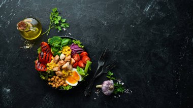 Buda kasesi: Tavuk, nohut, brokoli, domates, yumurta, kırmızı biber, siyah arkaplanda siyah bir tabakta soğan. Üst Manzara. Metnin için boş yer.