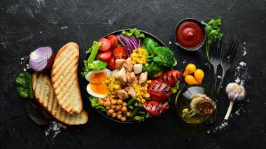 Buda kasesi: Tavuk, nohut, brokoli, domates, yumurta, kırmızı biber, siyah arkaplanda siyah bir tabakta soğan. Üst Manzara. Metnin için boş yer.
