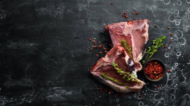 Kemiğin üzerinde çiğ iki biftek ve siyah taşlı bir arka planda baharatlar. Üst Manzara. Kırsal biçim.