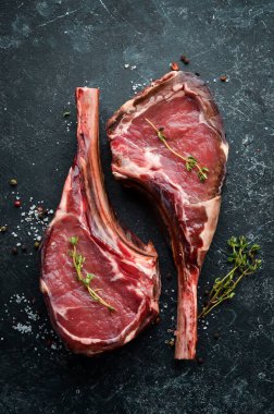 Kemiğe baharatlı iki çiğ biftek. Siyah taştan bir arka planda. Üst Manzara. Boş kopyalama alanı.