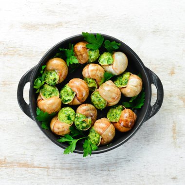 Beyaz ahşap arka planda siyah bir tabakta sarımsaklı tereyağlı fırında salyangoz ve taze otlar. Üst Manzara. Metnin için boş yer.