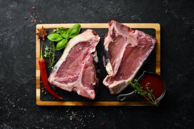 Kemiğin üzerinde çiğ biftek, ahşap tahtada baharatlar ve otlar. Üst Manzara. Siyah taştan bir arka planda.