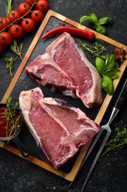 Kemiğin üzerinde çiğ iki biftek ve siyah taşlı bir arka planda baharatlar. Üst Manzara. Kırsal biçim.