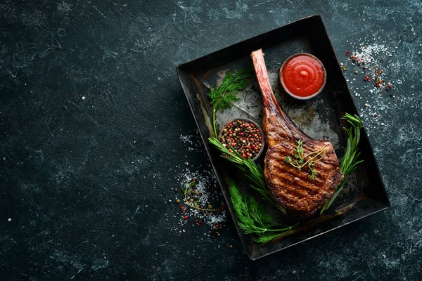 Grilled steak on the bone. Tomahawk steak on a black stone background. Top view. Free copy space.