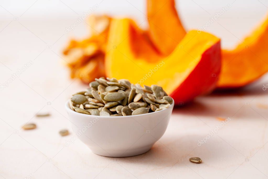 Peeled pumpkin seeds in a bowl with pieces of sliced fresh pumpkin on a wooden light background. Copy space text.