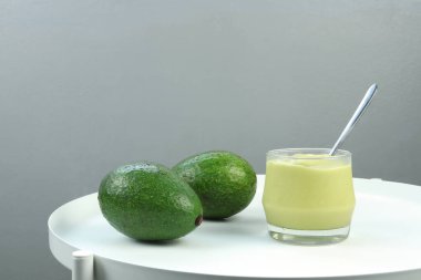 Bir bardak taze avokado suyu, organik meyve kafesinde sergilenen organik avokadolarla birlikte beyaz masanın üzerinde sergilenmektedir. Arka planı gri beton kaplama, ferahlatıcı lif ve enerji sağlığı açısından zengin.