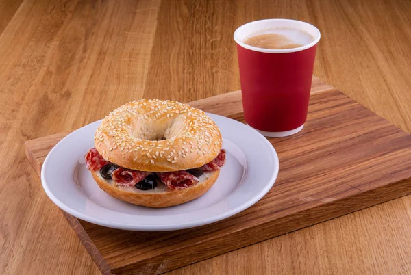 Bir fincan kahve ile ahşap bir tahtada beyaz tabakta salam ve zeytin sandviç