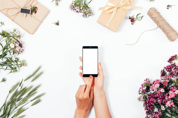 Female holding smart phone in hands among gifts and summer flowers, top view. Flat lay festive composition woman is touching screen of mobile device with her forefinger.