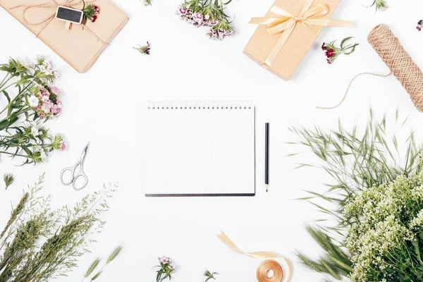 Top view of blank notepad among summer field flowers and gifts on white background. Flat lay floral mock-up with space for text on the empty sheet of diary.
