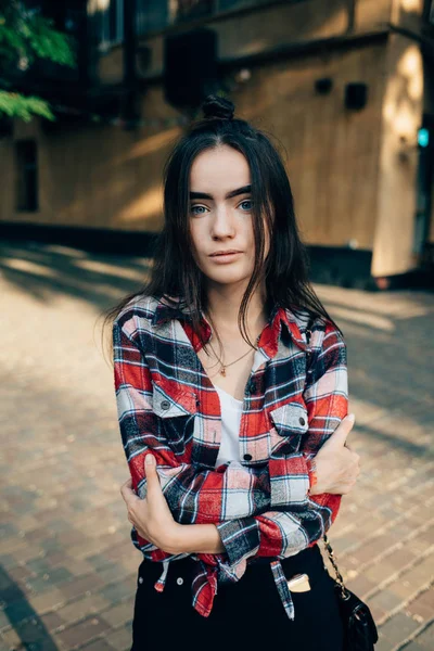 Teen girl with long dark hair looks at camera standing in city with green trees at summer day. Vertical portrait of young female wearing plaid red shirt hugging herself.