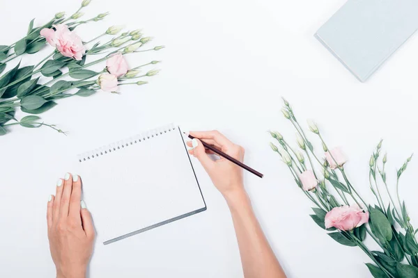 Woman\'s hand with pencil making notes on blank sheet near book and bouquet of flowers, top view. Flat lay composition stylized feminine workplace.