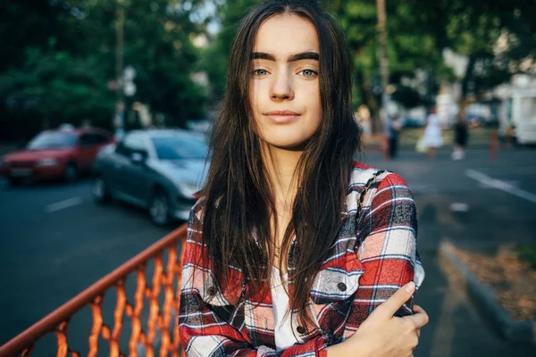 Teen girl with long dark hair wearing plaid shirt looks at the camera and smiles. Portrait of confident happy young woman standing in city enjoying of summer day.