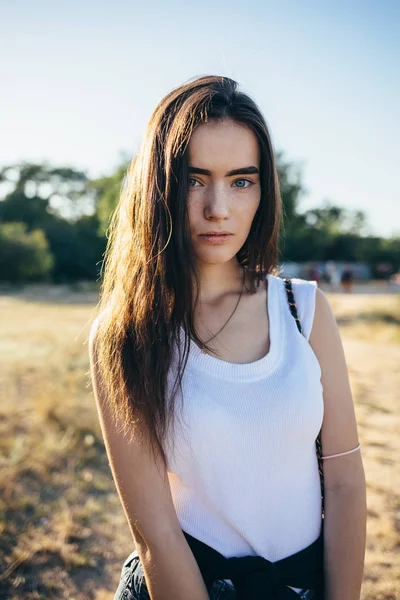 Close-up candid portrait of serious young woman with long brown hair on the background of summer meadow the sun shines from behind, vertical framing.