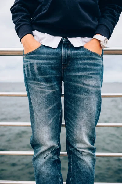 Details of casual clothing in blue and white colors. Lifestyle close-up of fashionable female wearing wristwatches holding hands in pockets of jeans standing near sea, vertical framing.