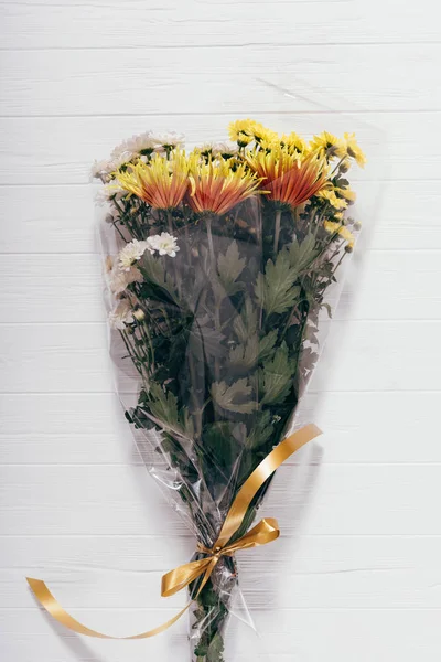 Lush long bouquet of yellow and orange asters packed in transparent cellophane with bow. Vertical image of fresh gift flowers on white wooden table, top view.