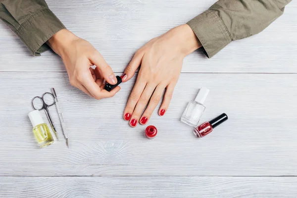 Girl's hands apply red polish on nails next to scissors, scraper, bottles of glitter lacquer, base and moisturizing oil on white table, top view. Young woman does manicure to herself.