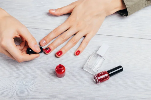 Girl\'s hands apply red polish on nails next to bottles of lacquer and base on white table, top view. Close-up young woman does manicure to herself.