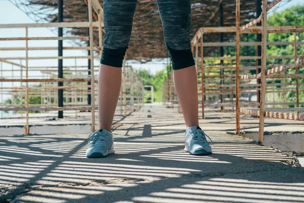 Fitness sporty woman in sportswear outdoor on morning, ready to exercise. Close up of female legs in short gray leggings standing over old building background at sunny summer day.