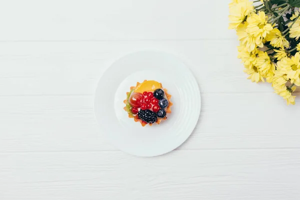 Bright small berry cake and bouquet of flowers on white wooden table, top view. Flat layout delicious fruit tartlet dessert on plate.