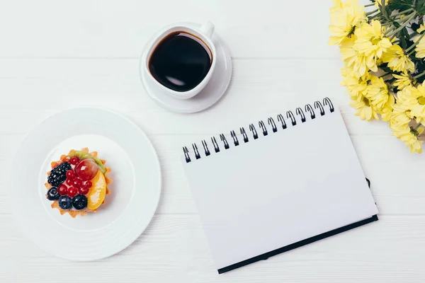 Open blank notepad next to cup of strong coffee, small berry cake and flowers on white wooden table, top view. Festive floral flat lay of feminine workplace at break.