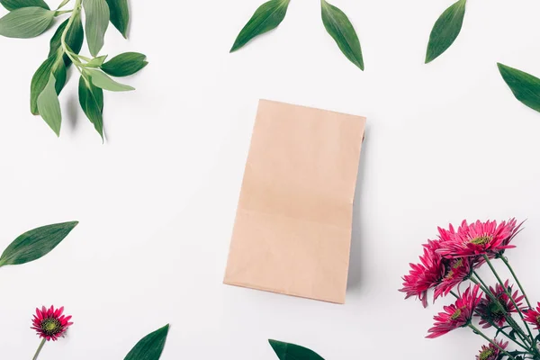 Brown paper bag in center of flat lay floral frame made of fresh green leaves and blooming pink flowers on white background, top view.