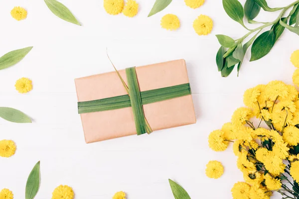 Natural gift box packaging among flat lay composition of yellow flowers and green leaves on white wooden table, view from above.