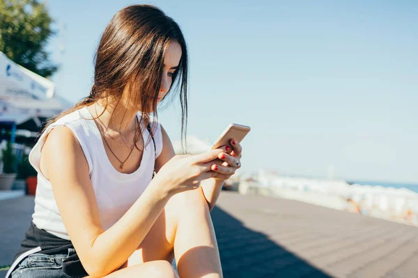 Young woman using smart phone outside. Female wearing white top sitting on summer sea coast looking at mobile device.