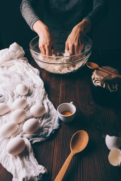 Female\'s hands kneading dough in big bowl on brown wooden kitchen table near eggs and jar of jam. Process of cooking homemade cookies.