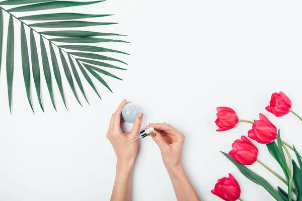 Female\'s hands with gold manicure apply cosmetic serum from bottle using pipette next to palm leaf and bouquet of flowers on white table, top view.