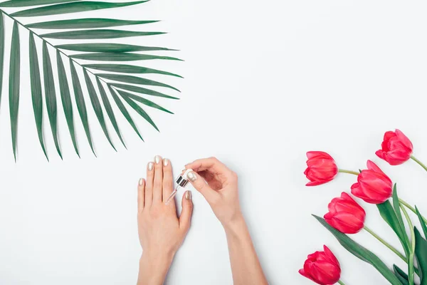 Female\'s hands with gold manicure apply cosmetic serum on skin using pipette next to palm leaf and bouquet of flowers on white table, top view.