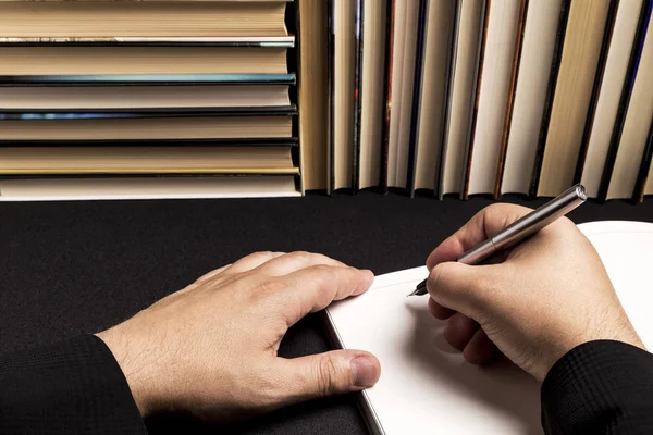 the hand of a man writing in a blank book beside a book background