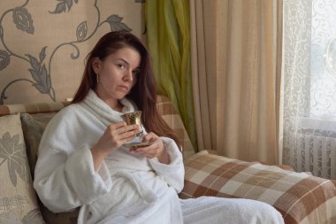 Beyaz bornozlu bir kız kanepede dinleniyor ve çay içiyor..